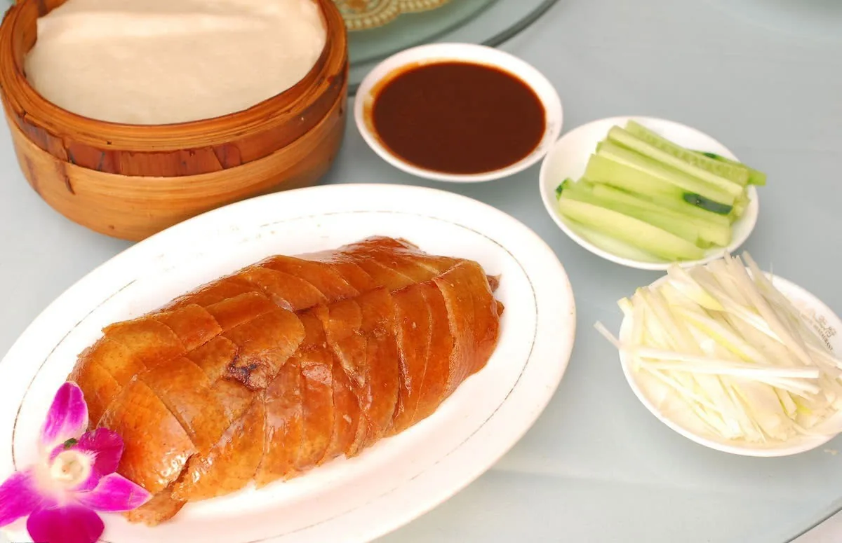 A roast duck on a white plate with pancakes, sauce and sliced cucumber