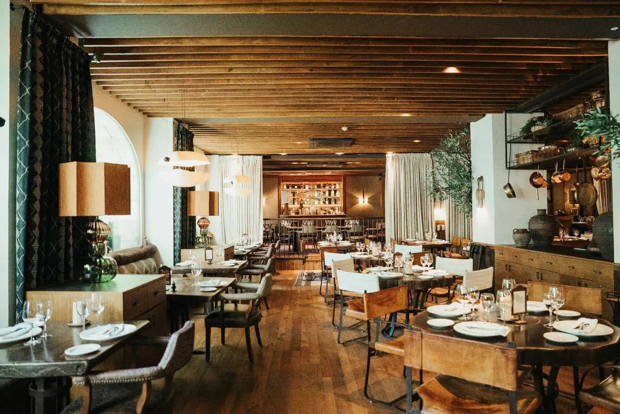 A large dining room with a slatted timber ceiling, arched window behind patterned drapes, cream globe pendants and table lamps, round live-edge tables laid with white plates and wine glasses, a lit back bar at the far end and copper pans hanging from a rack on the right.