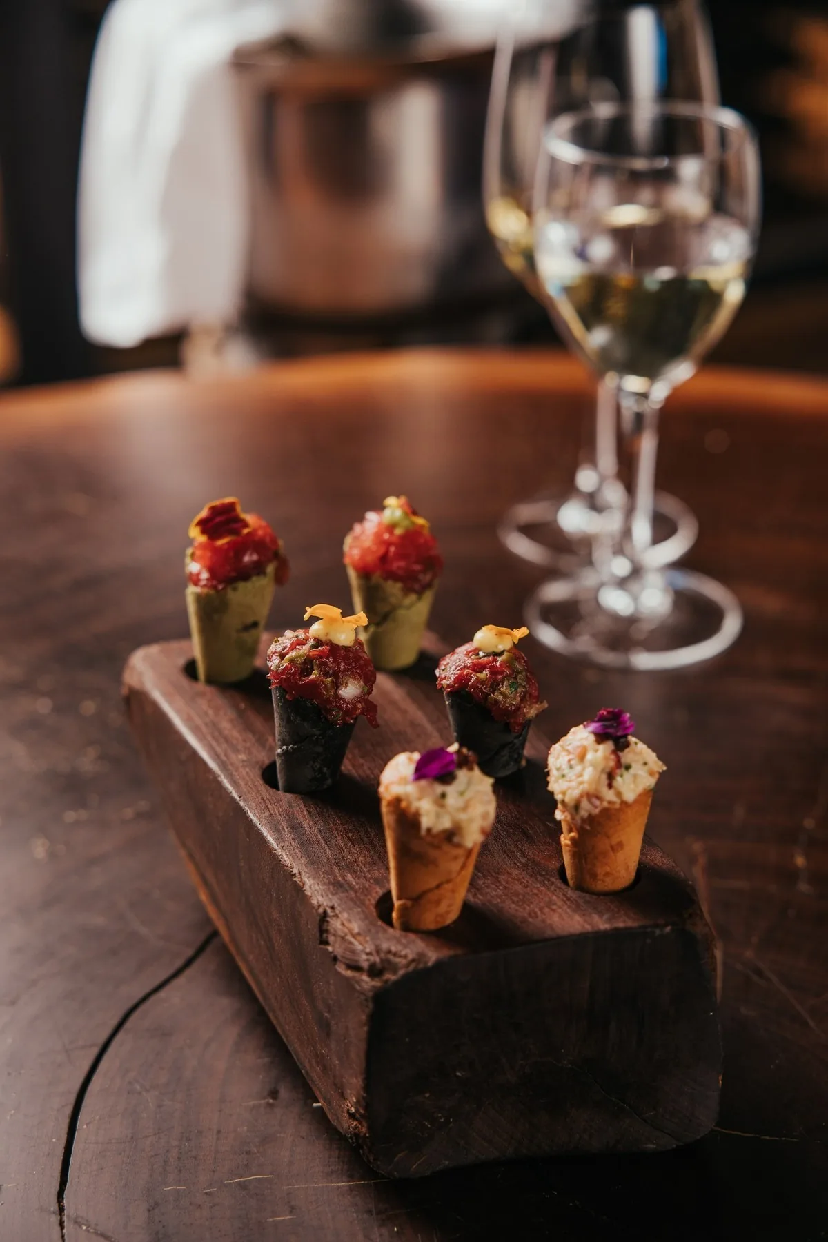 Small filled cones of appetisers on a wooden block beside a glass of white wine