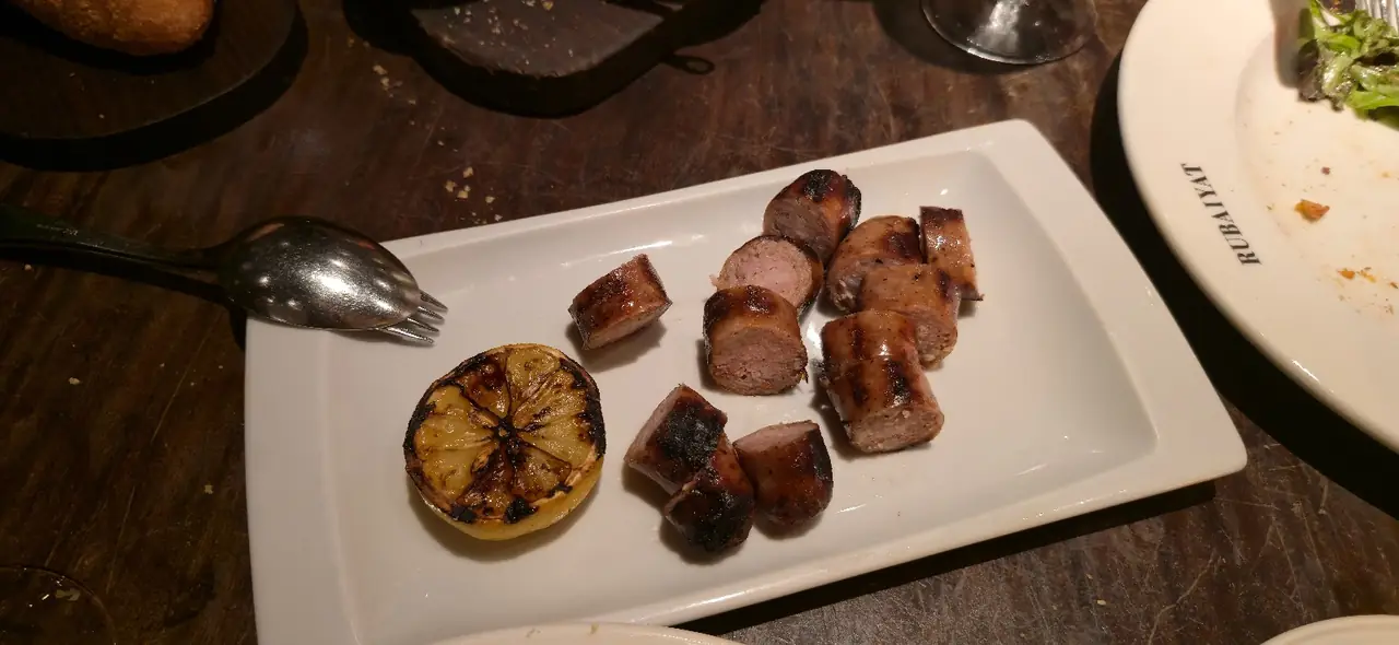 Grilled sausage rounds with a charred lemon half on a white plate, Rubaiyat plate visible