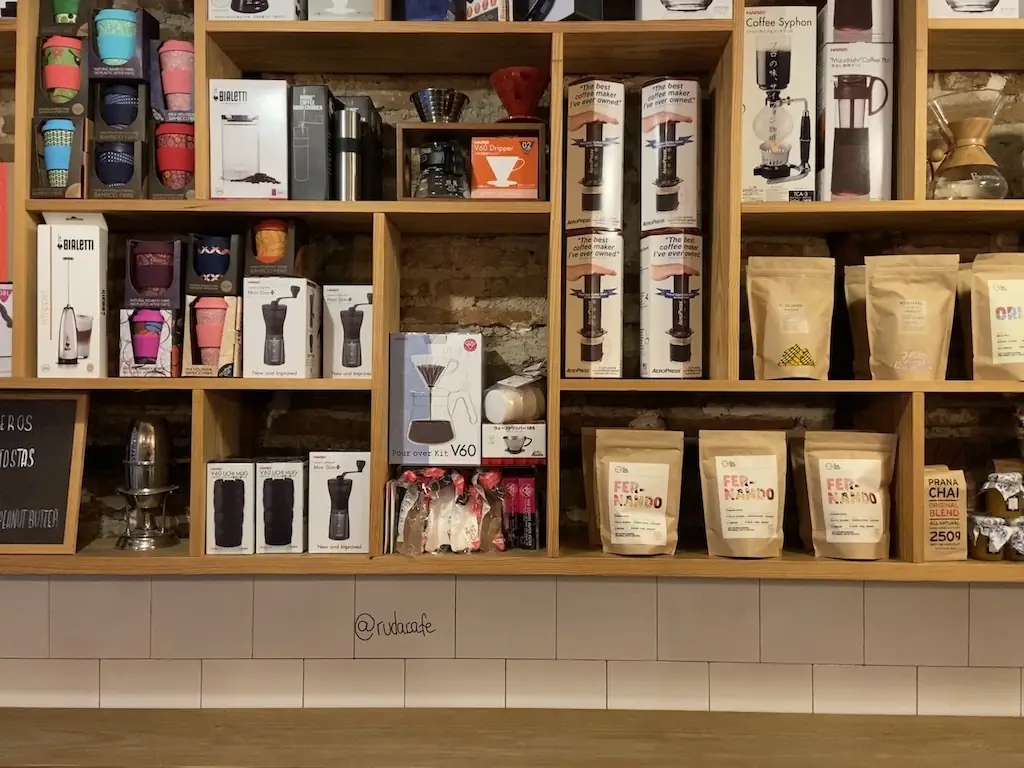 Cafe interior wooden shelving stocked with coffee gear, brew kits and bags of coffee against an exposed brick wall, with @rudacafe handwritten on the tiled counter front