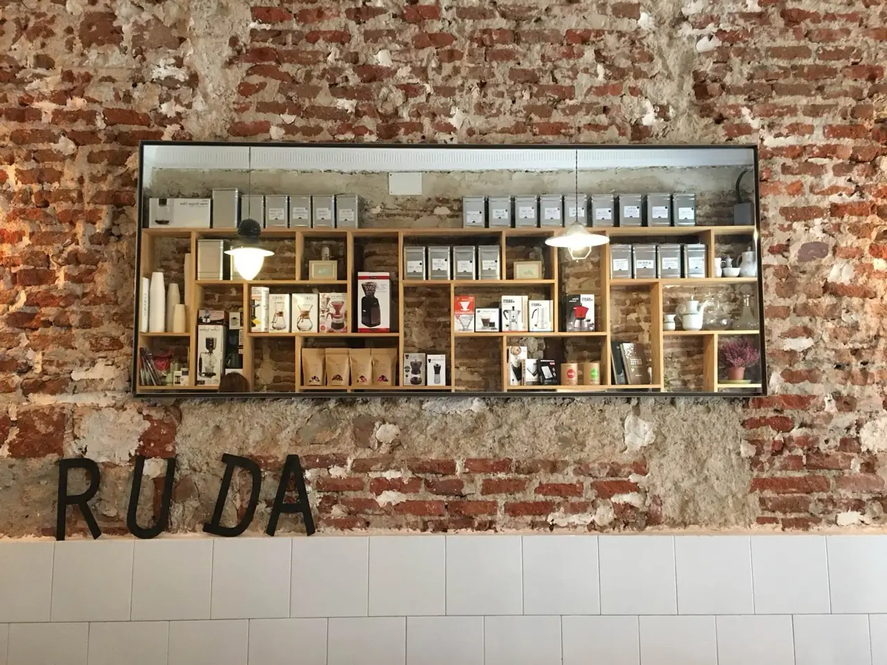 Cafe interior exposed brick wall with a glass-fronted display of coffee gear and tins, and dimensional black RUDA lettering below