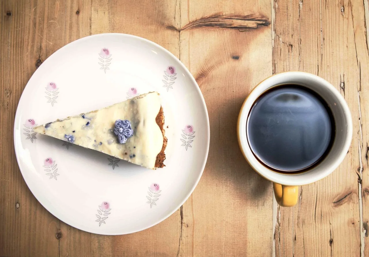 A slice of cake on a plate beside a black coffee in a cup on a wooden table, seen from above