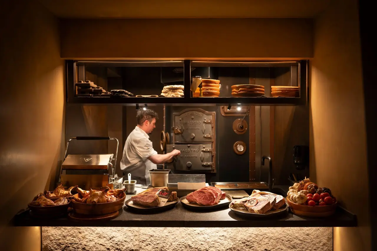 A lit service counter in front of an open kitchen: whole roast suckling pigs in glazed earthenware dishes, a dry-aged steak with a producer tag, a thick marbled ribeye on a metal plate, cut bone-in beef, and a bowl of tomatoes and garlic, with a cook in whites working at a riveted iron wood oven behind.