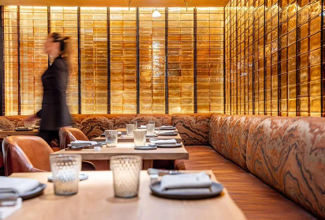 A dining corner backed by a floor-to-ceiling wall of amber glass bricks, with a long marbled orange banquette, pale wood tables set with grey plates, folded napkins and cut-glass tumblers, and a member of staff walking past in motion blur.