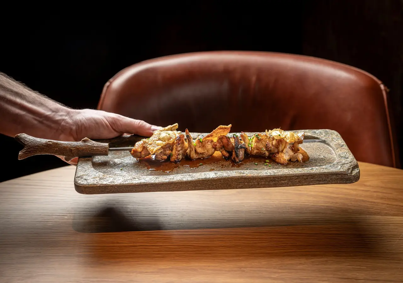 A hand holds out a rough stone slab by an antler handle, carrying a metal skewer threaded with charred grilled poultry pieces and mushrooms, glazed in dark jus with crisp golden wafers and chopped chives, a tan leather chair behind.