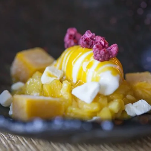 Dessert on a dark slate plate - diced mango and pineapple with a quenelle of cream, mango sauce, cubes of marshmallow and freeze-dried raspberries on top