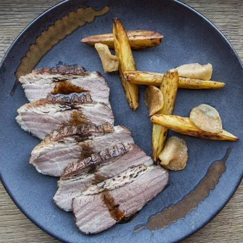 Slices of grilled pork fanned on a blue-grey plate with roast potato wedges, whole roasted garlic cloves and a drizzle of dark sauce