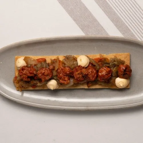 A long rectangular flatbread on an oval grey plate, topped with rounds of red sobrasada-style sausage, softened peppers and onions, and dots of aioli