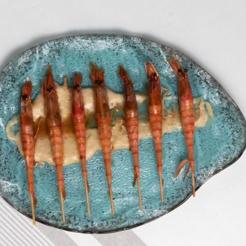 Seven whole red prawns on wooden skewers laid in a row over a pale sauce on a mottled turquoise ceramic plate