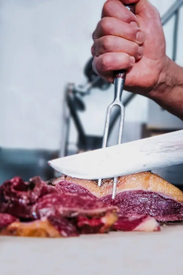 Hands carving grilled picanha beef with a long knife and a two-pronged fork over a cutting board