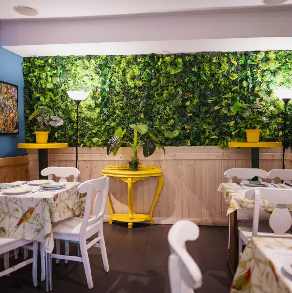 Restaurant dining room with white chairs, tables set with printed cloths, a green foliage wall and a yellow console table