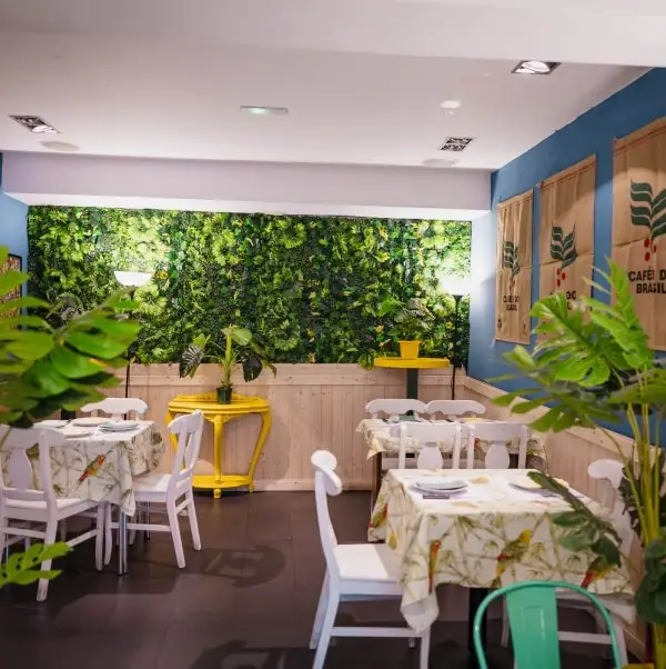 Wider view of the dining room with green foliage wall, coffee-sack decor, white tables and chairs and potted plants