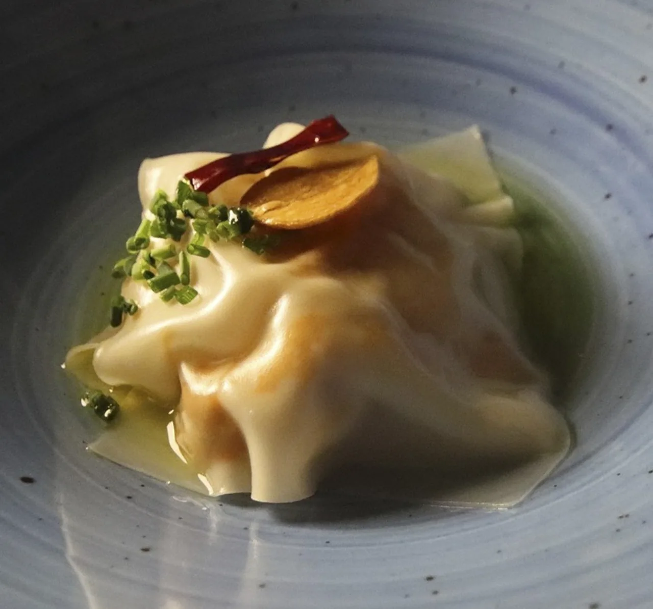 A folded pasta parcel in a clear broth with fresh herbs in a shallow blue-rimmed bowl