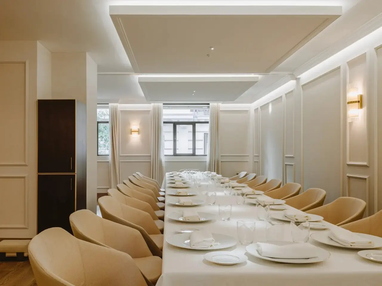 A long private dining table under a white cloth set with white plates, folded napkins and rows of glasses, lined with pale upholstered chairs in a cream panelled room