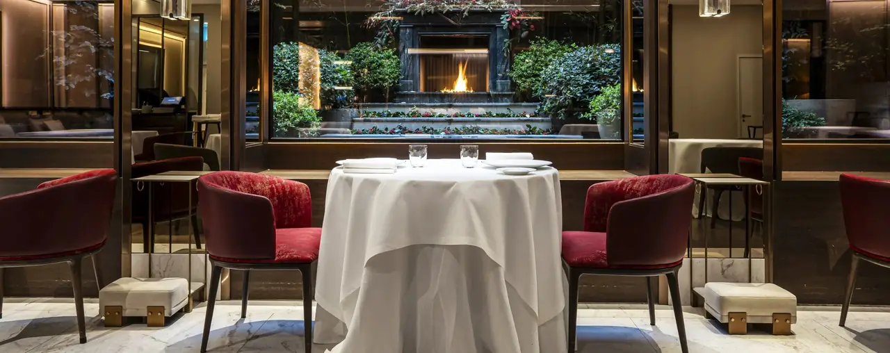 A table for two in a white cloth with red velvet chairs, set with plates and water glasses in front of a window looking onto a planted courtyard with a lit fireplace