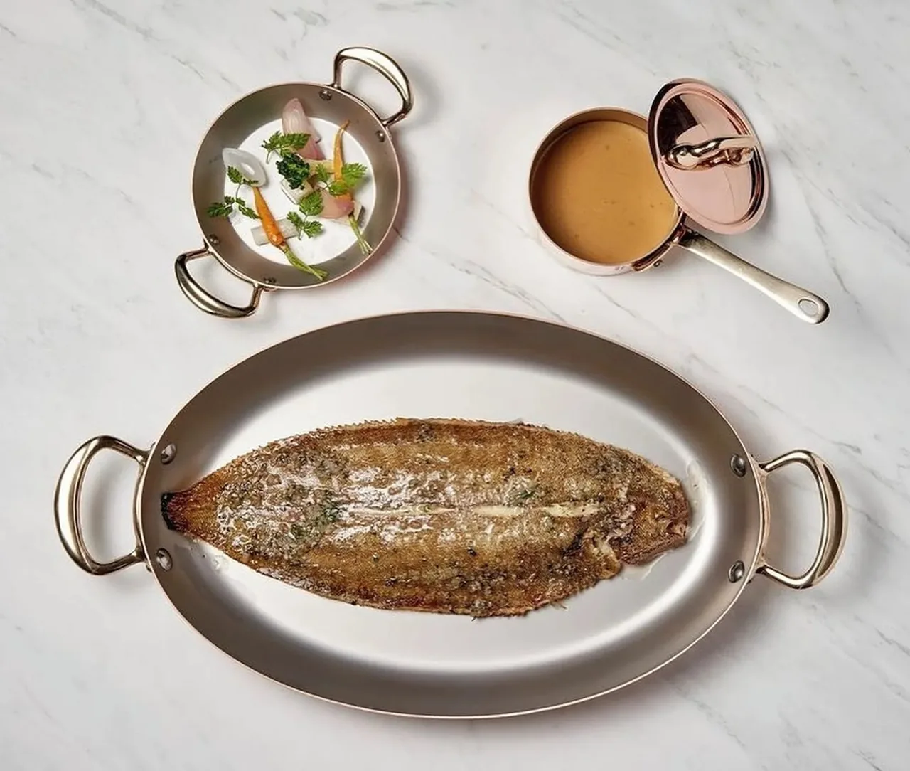 A whole flat fish in an oval metal pan with small copper pots of sauce on a marble table