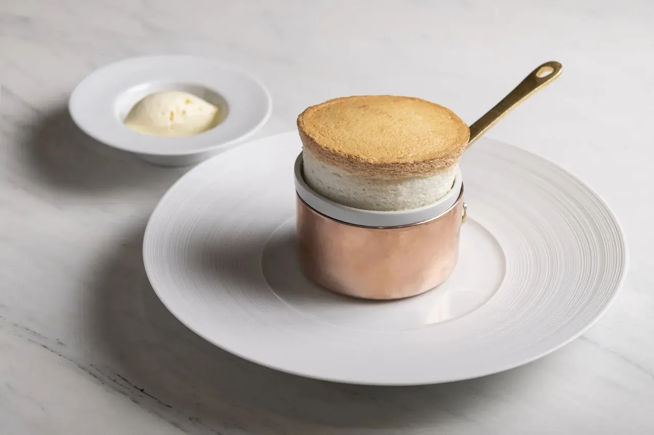 A puffed soufflé in a small copper pan on a white plate with a small dish of cream