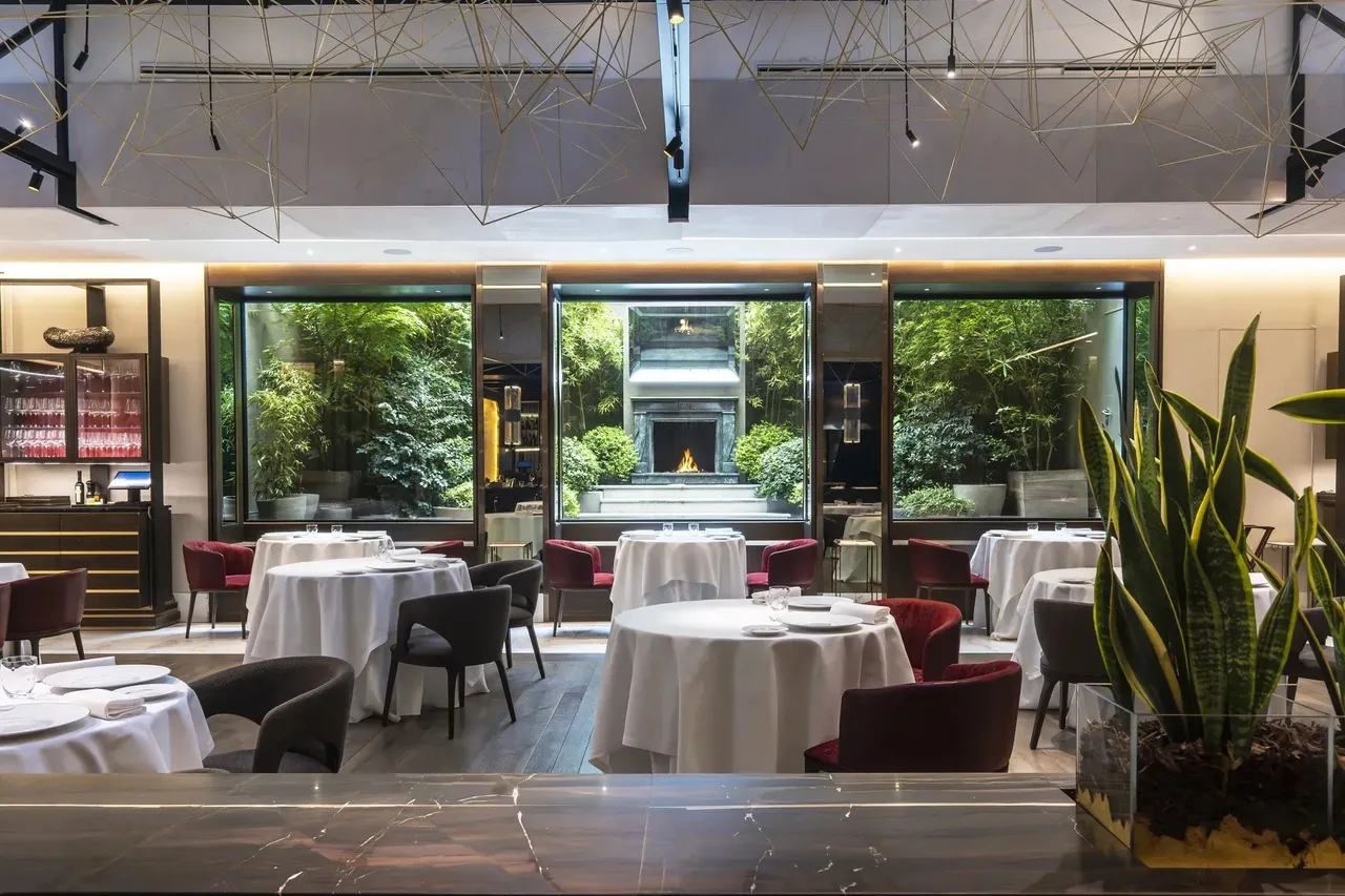 A dining room with white-clothed round tables and a glass wall opening onto a green courtyard