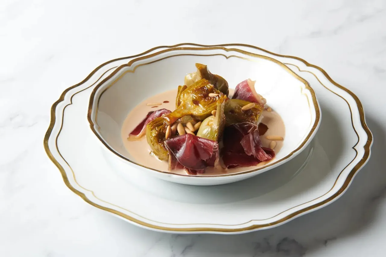 A gold-rimmed white bowl on a matching plate holding a plated dish with artichoke and dark leaves