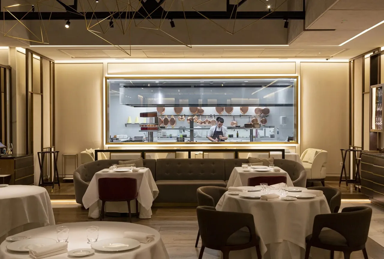 A dining room with white-clothed tables, an open kitchen behind glass and warm lighting