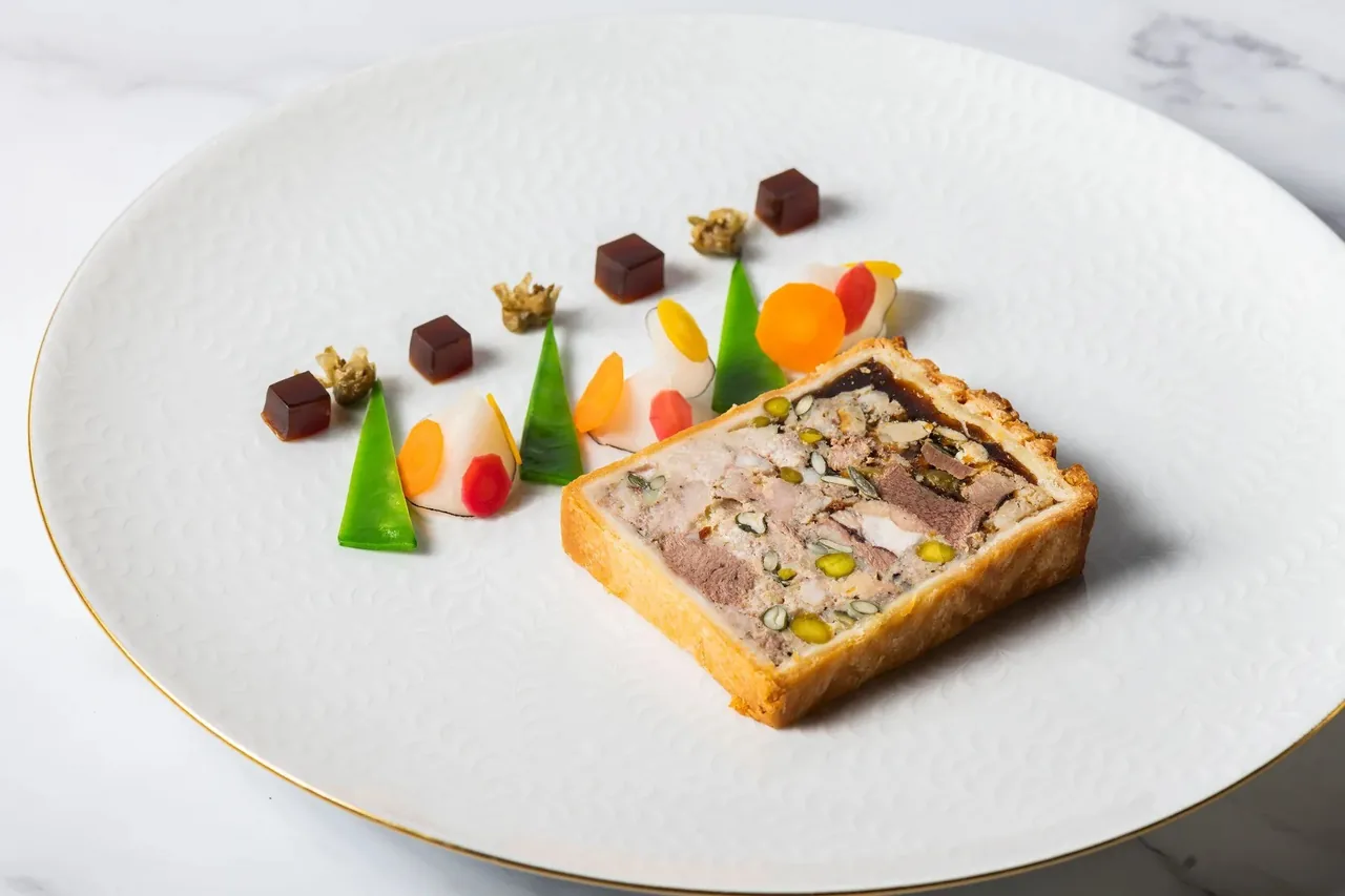 A large white plate with a slice of terrine in pastry and small colourful vegetable garnishes