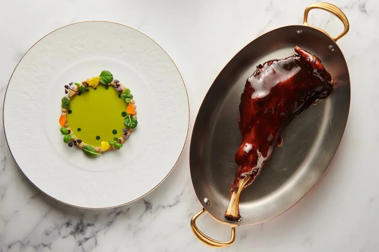 A round white plate with a green purée and a copper pan holding a glazed meat shank