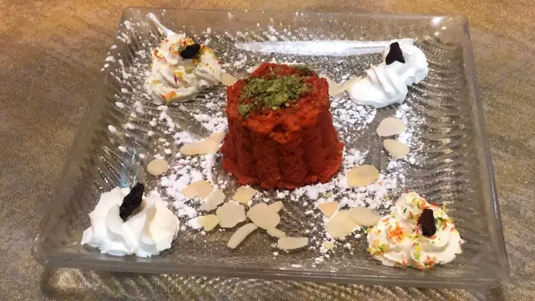 Molded red carrot halwa dessert topped with green pistachio, on a glass plate dusted with icing sugar and dotted with whipped cream and sliced almonds