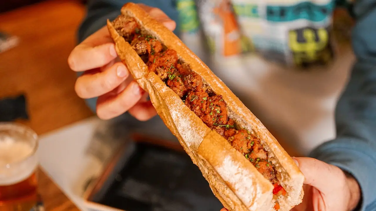 Crusty baguette sandwich packed with breaded, herb-flecked meat, held open in two hands, Sagàs Pagesos i Cuiners