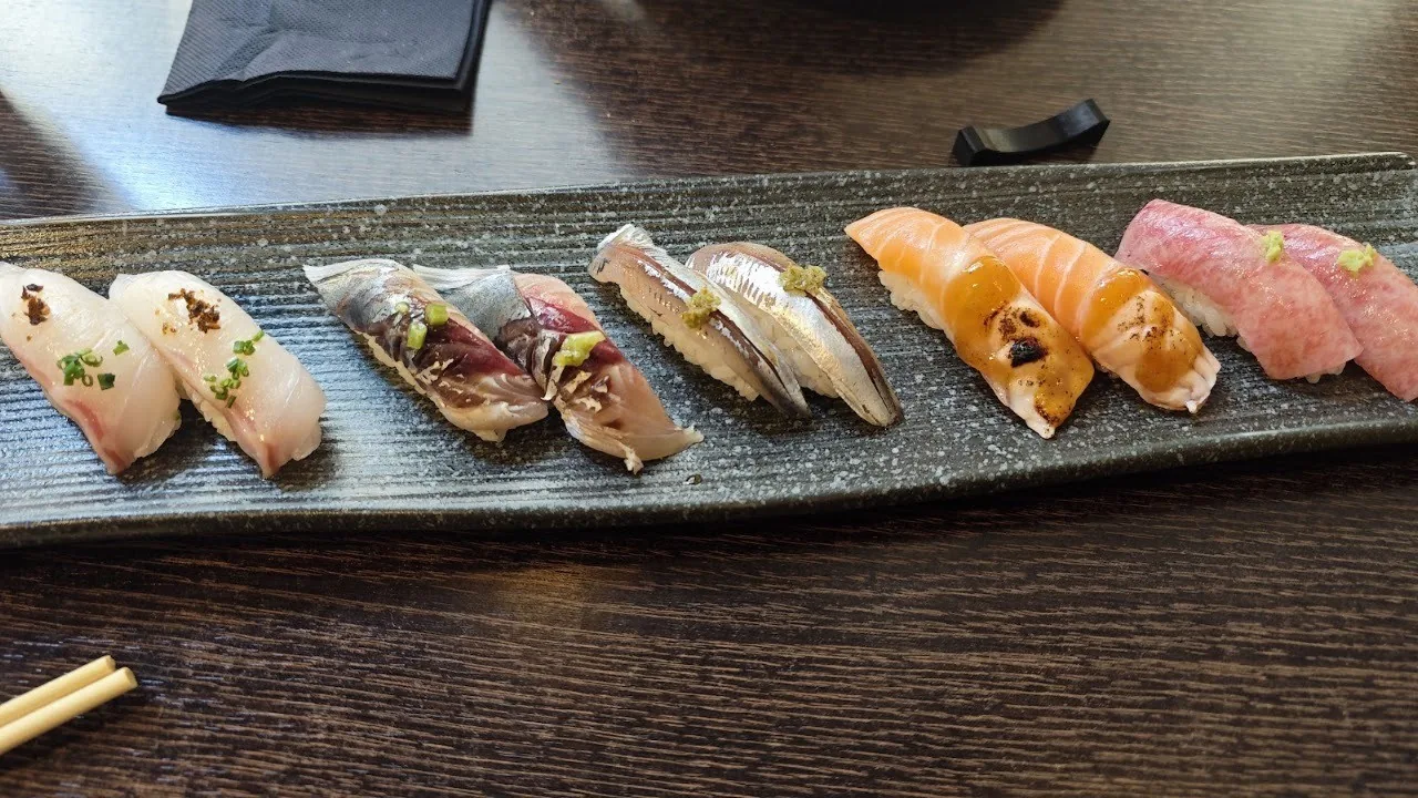 A row of nigiri on a long dark plate beside chopsticks