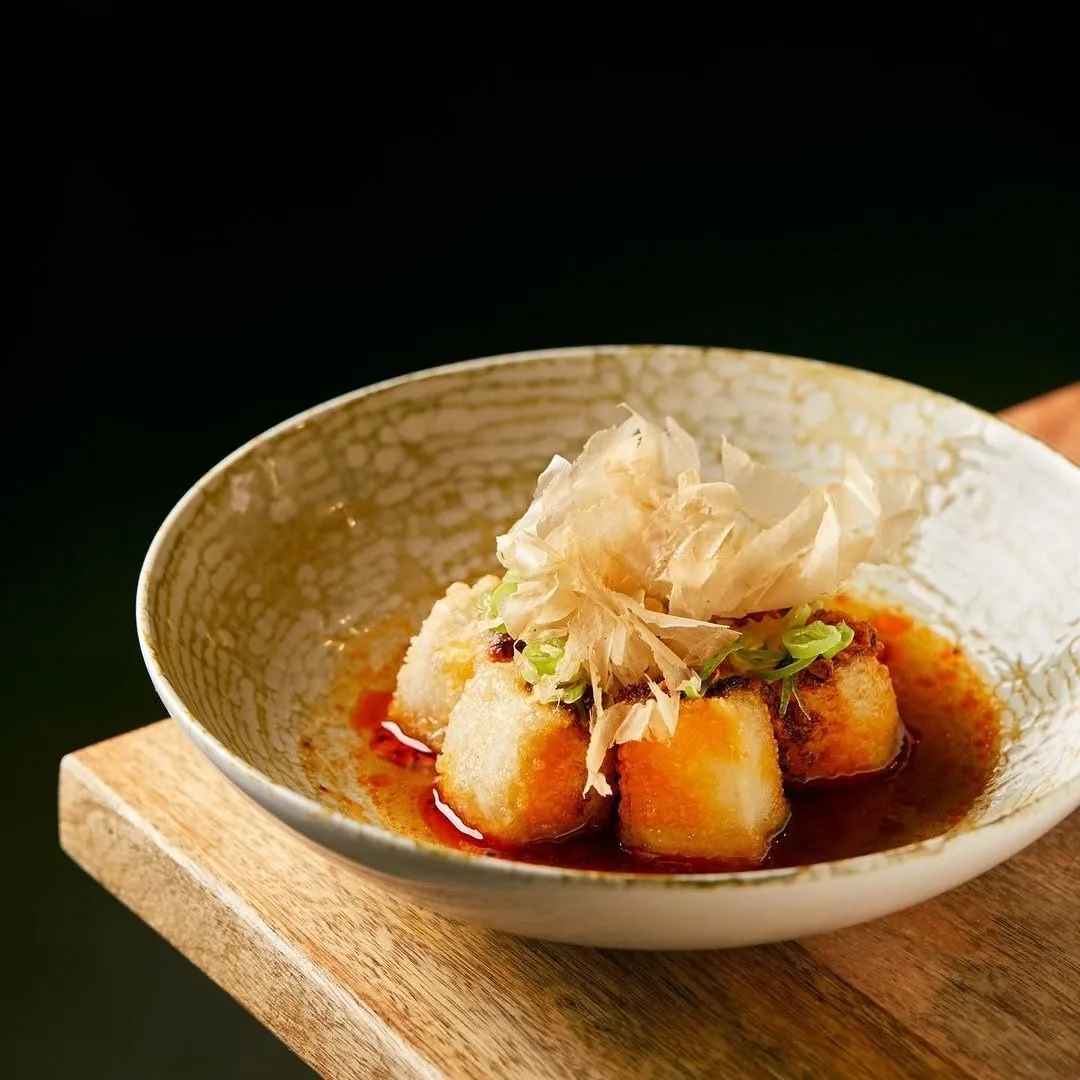 Fried tofu cubes in a red sauce topped with fine shavings in a shallow bowl