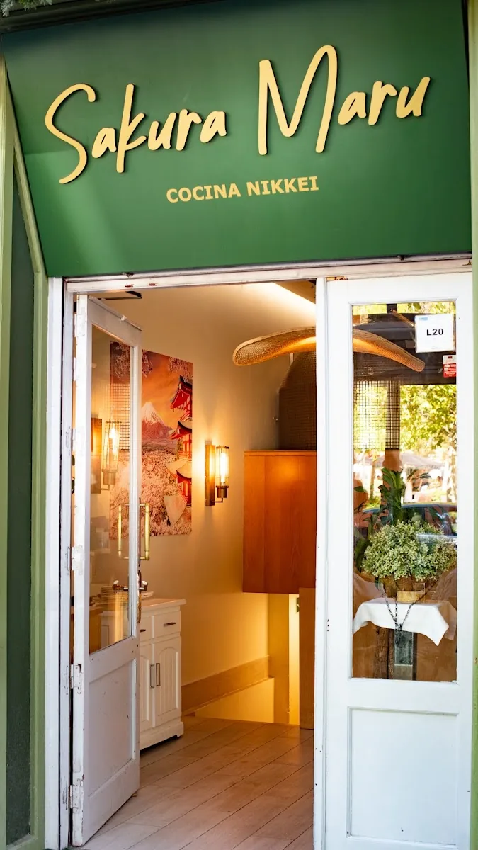 Sakura Maru's entrance under a green awning lettered in gold script with 'Cocina Nikkei', white double doors open onto a warm-lit hallway hung with a Japanese landscape poster