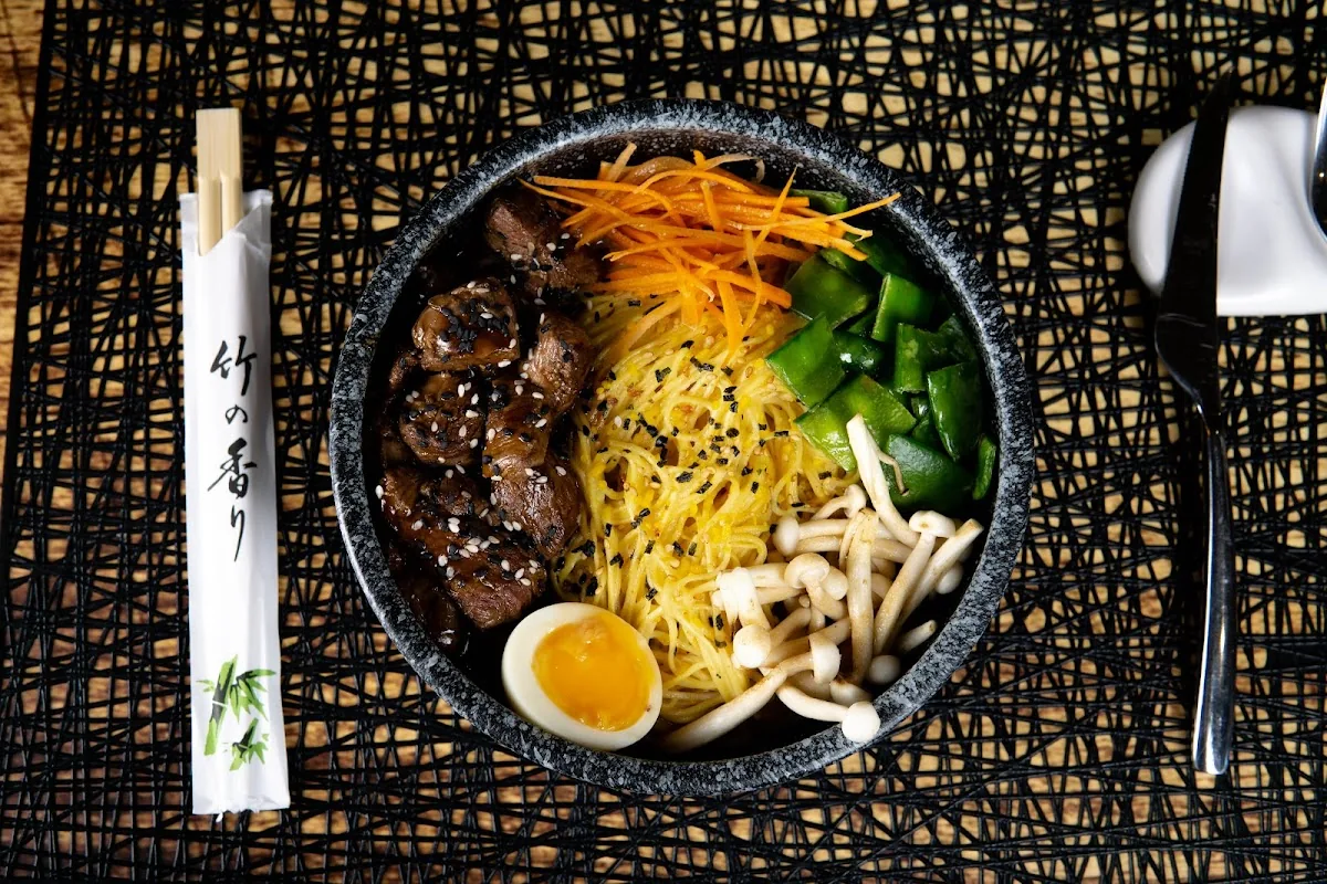 A stone bowl of egg noodles topped with sesame-glazed beef, julienned carrot, mangetout, enoki mushrooms and a soft-boiled egg, chopsticks alongside on a black lattice mat
