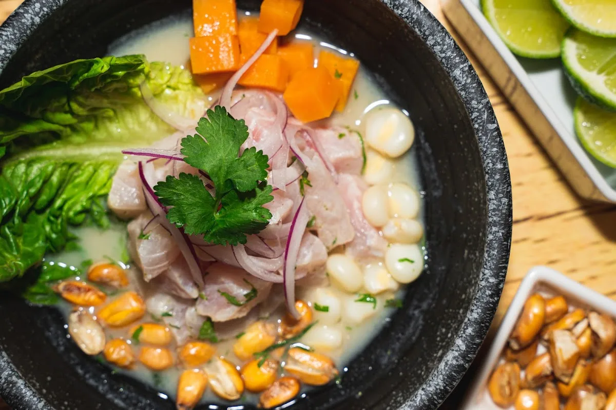 Ceviche clasico in a dark stone bowl, white fish in milky leche de tigre with red onion, cilantro, choclo corn, toasted cancha and sweet potato, lime wedges in a dish beside it