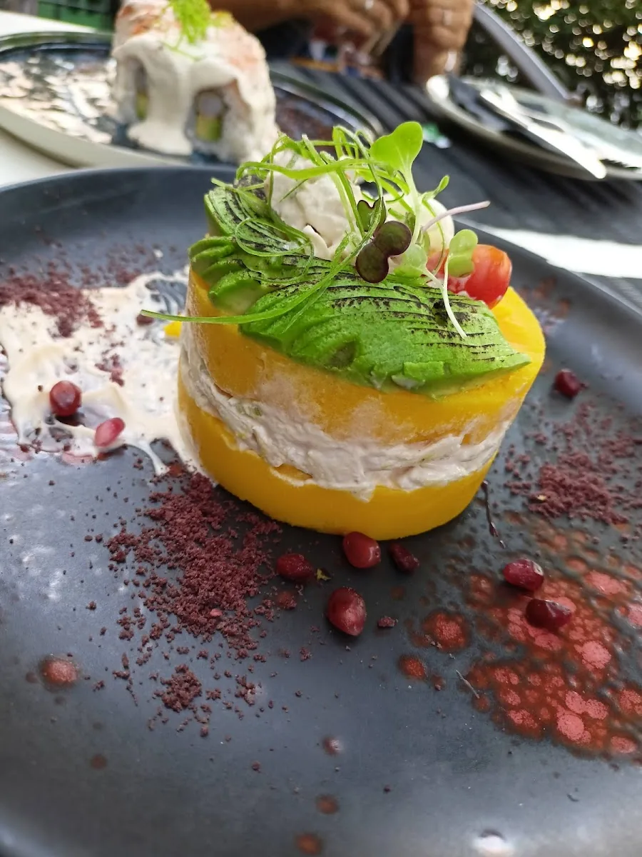 A causa limena on a slate plate, a round of yellow potato layered with a creamy filling, fanned avocado and microgreens on top, pomegranate seeds and purple powder scattered around