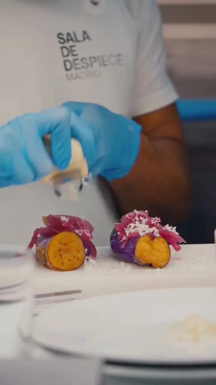 Purple-skinned potatoes topped with pickled red onion and grated cheese, plated with a chef finishing the dish.