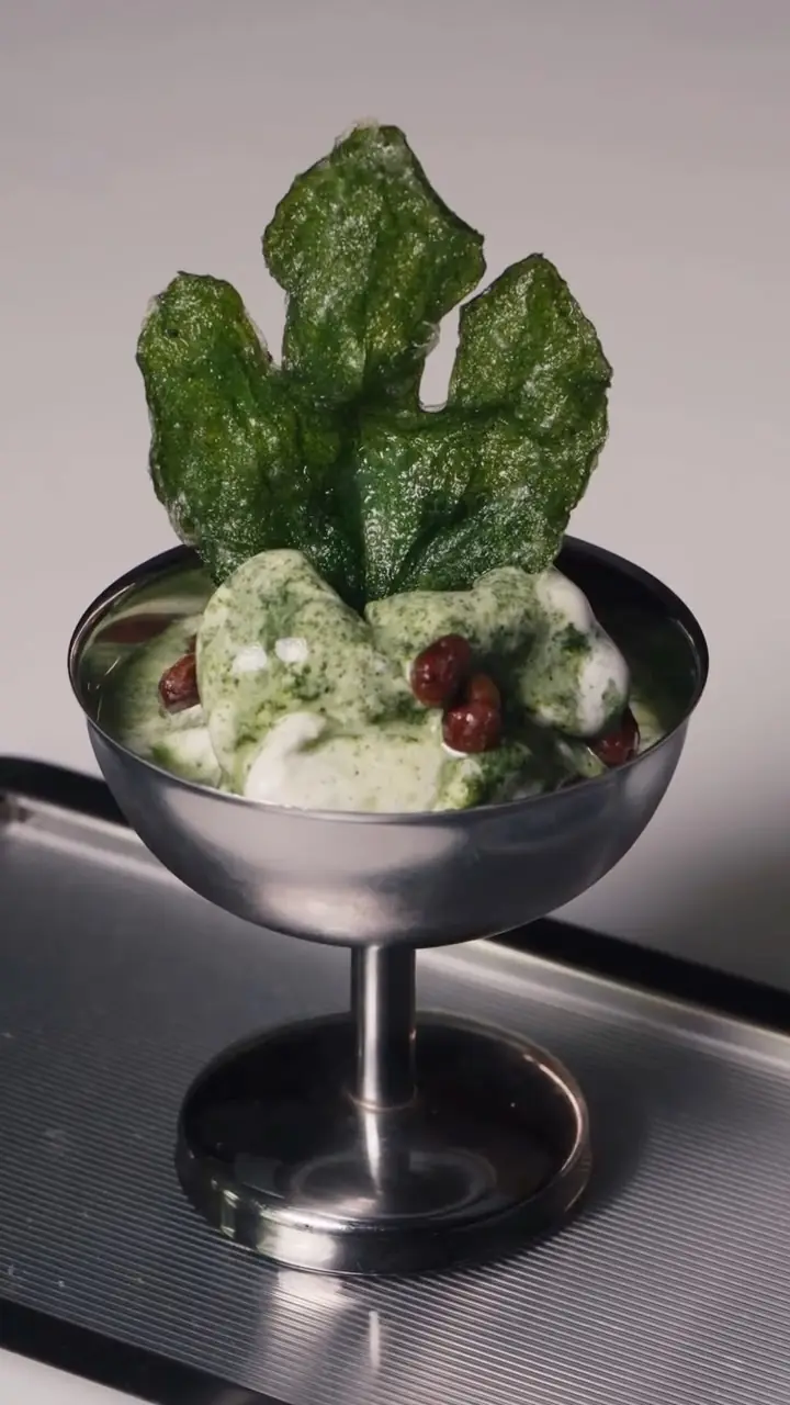 A fried spinach leaf and creamy green sauce with dark berries served in a stemmed metal cup.