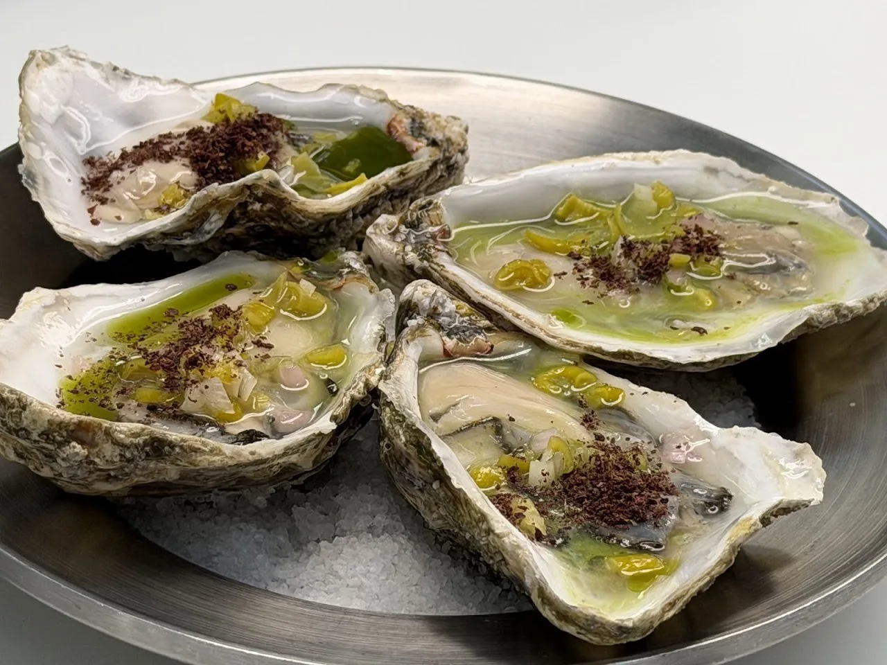 Four opened oysters with green dressing and dark crumbs on a metal plate