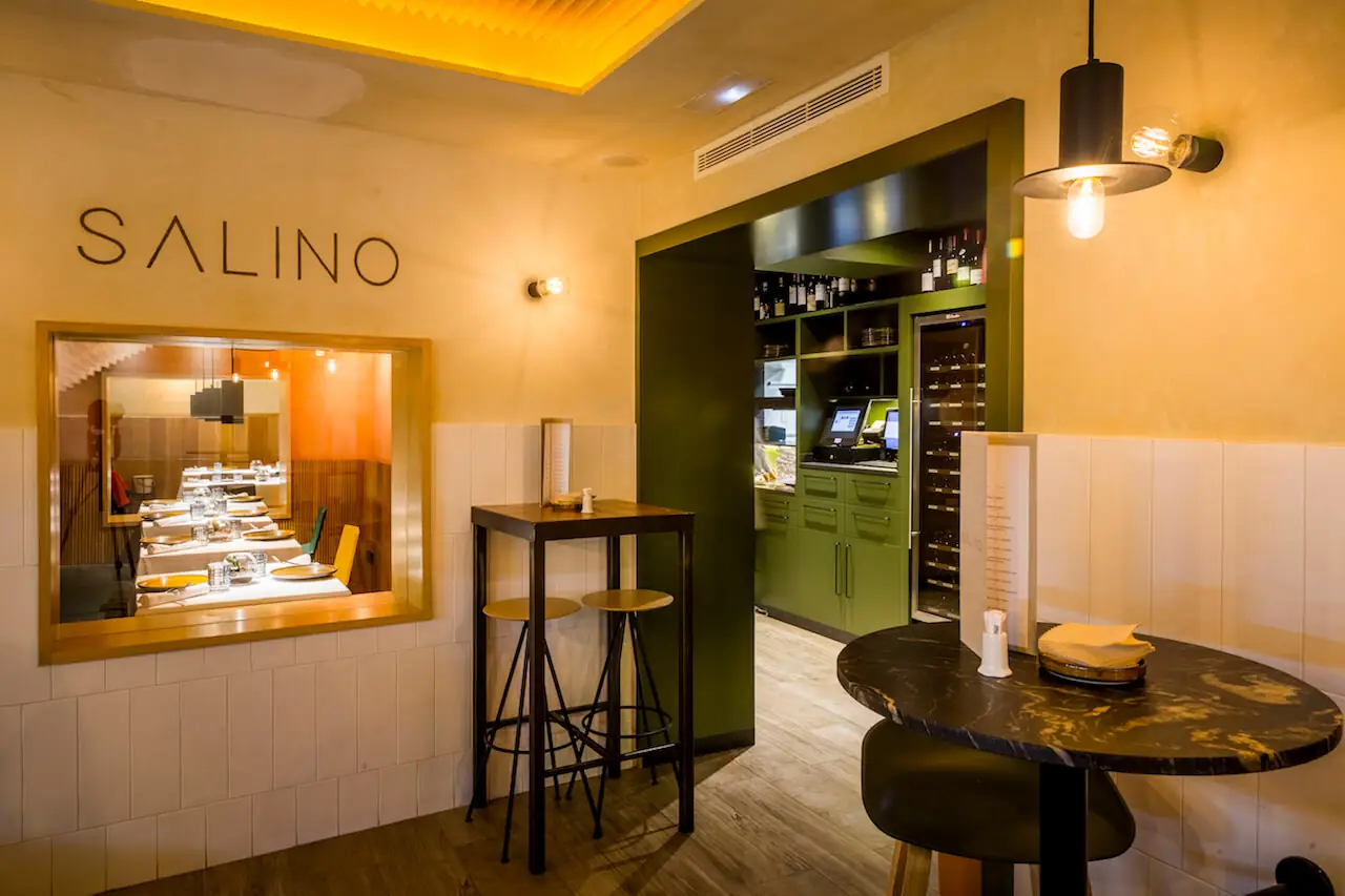 Salino interior corner showing the SALINO name on the wall, a framed window into the dining room, high stools and a green wine cabinet