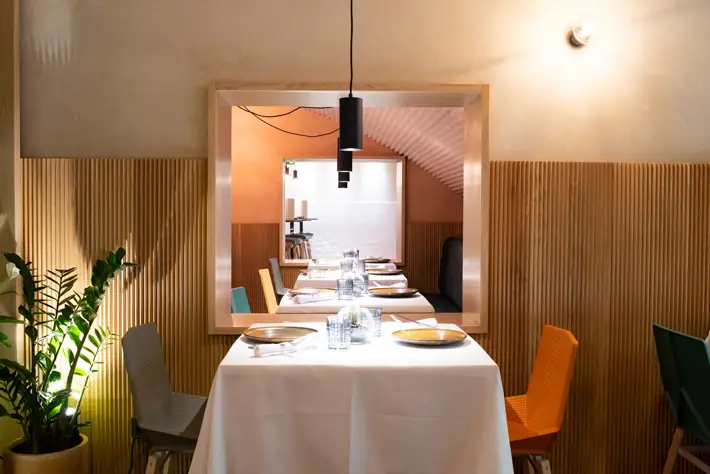 Intimate dining nook with a white-clothed table, colorful chairs, wood-slat walls, a potted plant and a framed interior window