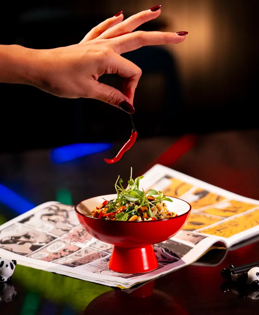Noodle-and-herb dish in a red pedestal bowl set on a comic-strip page, a manicured hand dropping a red chili from above