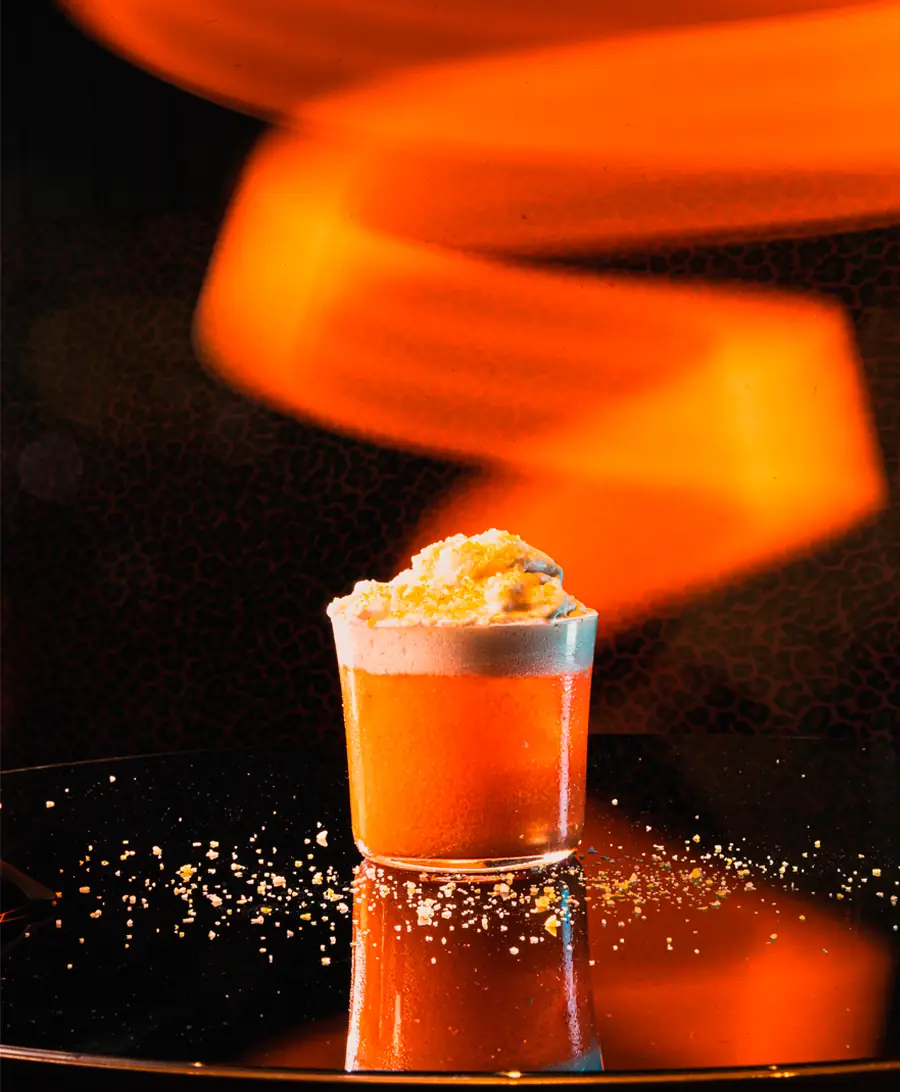 Frothy orange cocktail in a small tumbler on a dark reflective table lit by a swirl of orange light, scattered crumbs around it