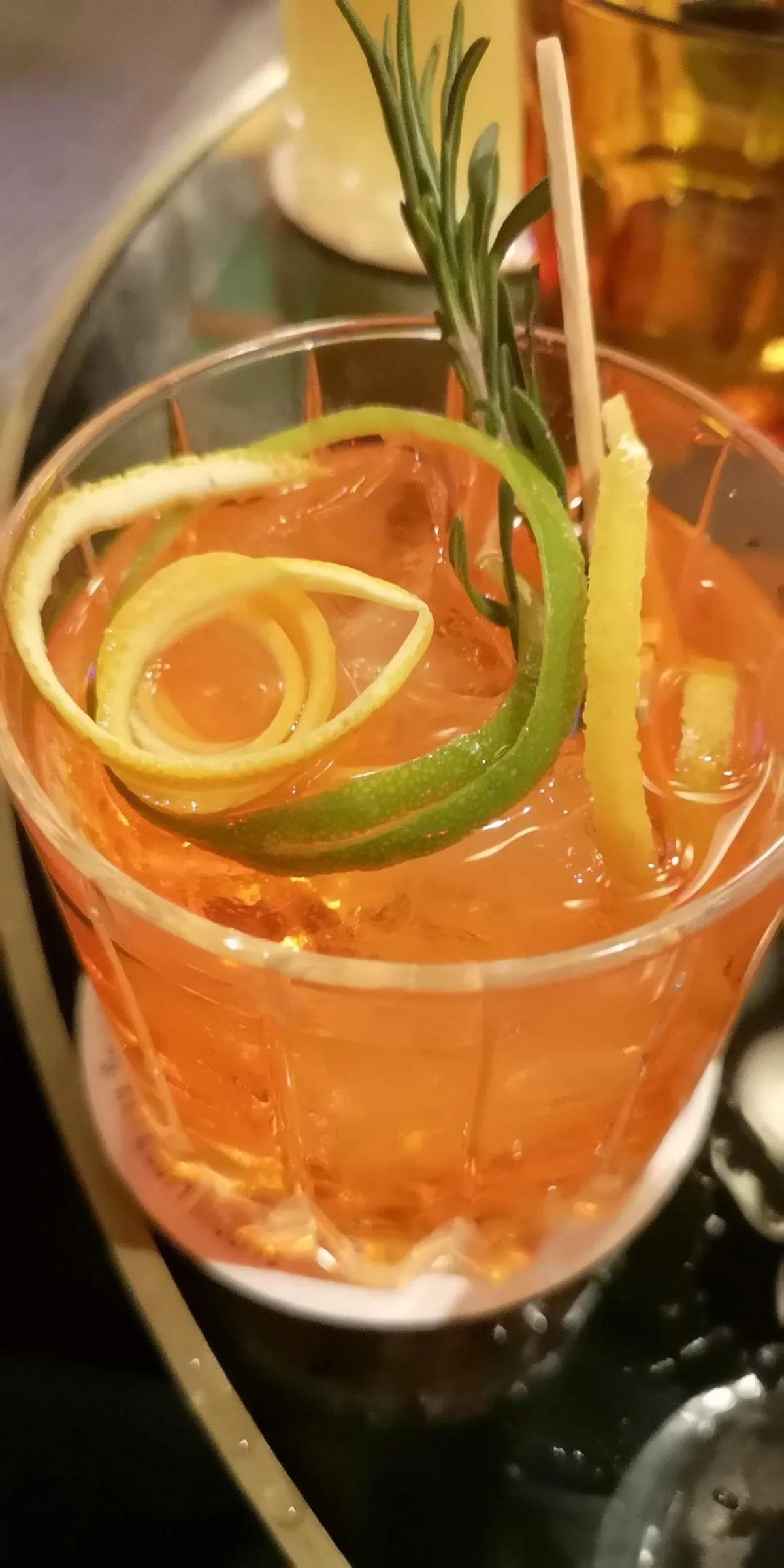 A tall orange cocktail with a rosemary sprig and citrus peel in a glass