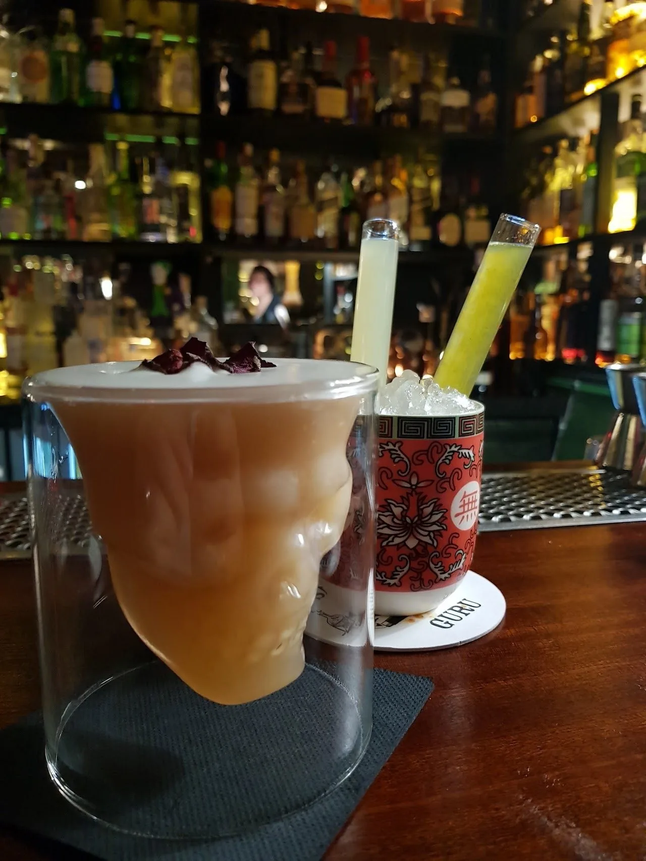 A cocktail in a skull-shaped glass on a wooden bar with bottles behind it