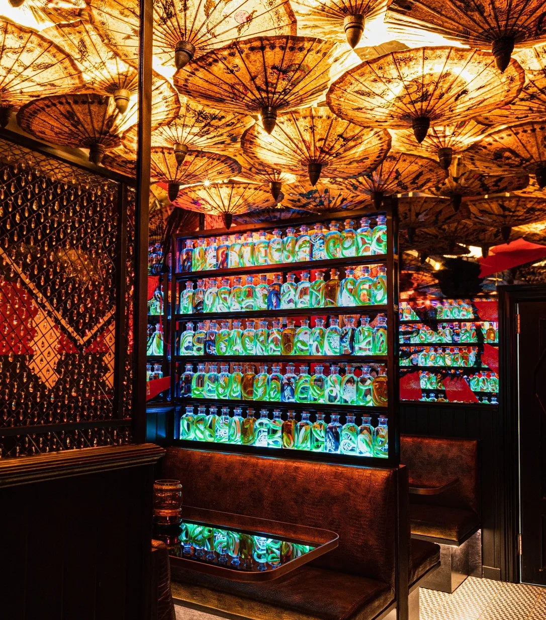 A leather booth beside a lit shelf of green glowing bottles under an ornate ceiling
