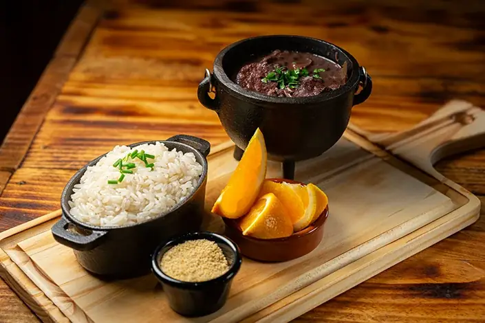 Feijoada served in a small cast-iron pot with white rice, farofa, and orange wedges on a wooden board, Samambahia Restaurante & Cachaça Bar