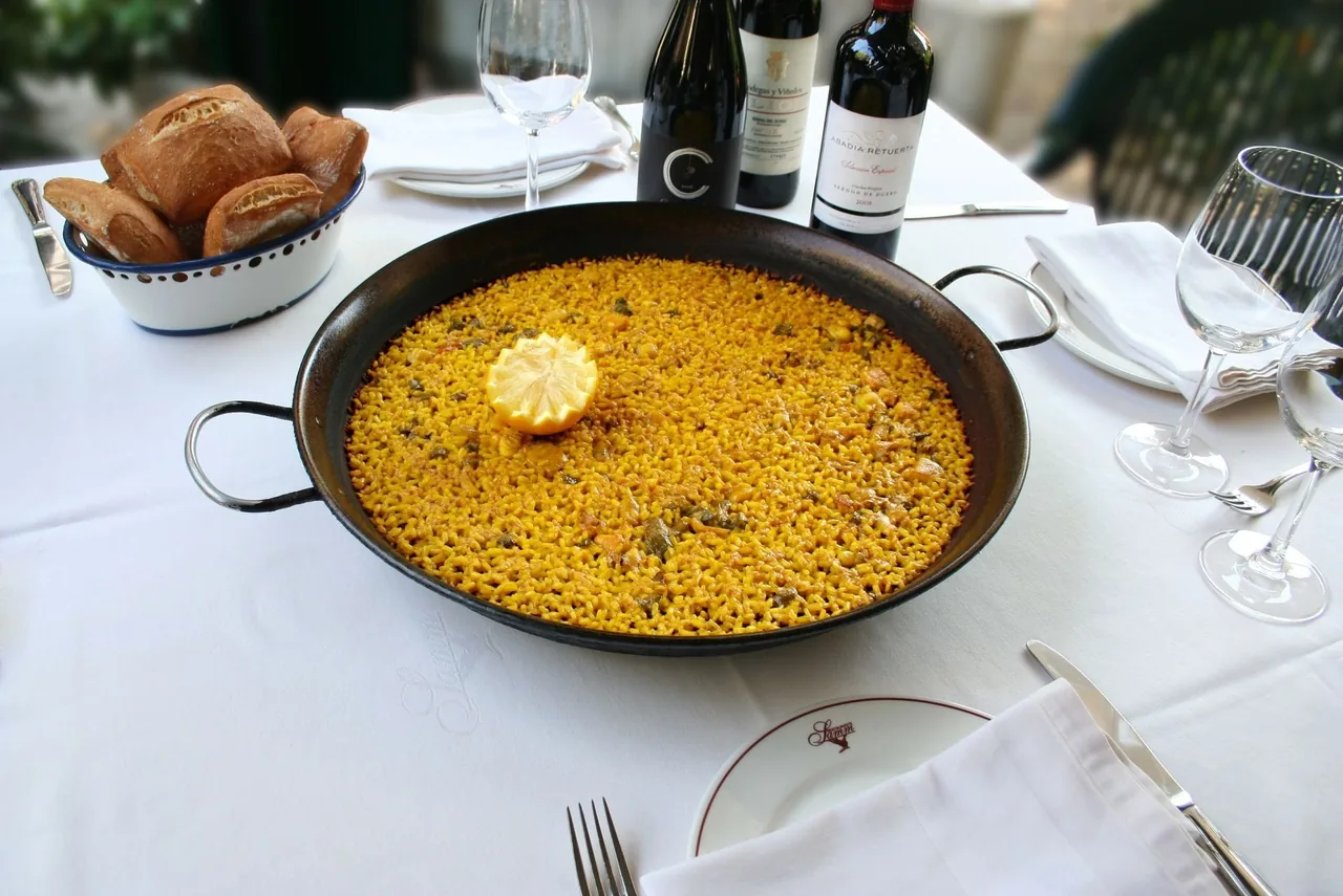A large black paella pan of golden rice with a lemon in the centre on a white tablecloth