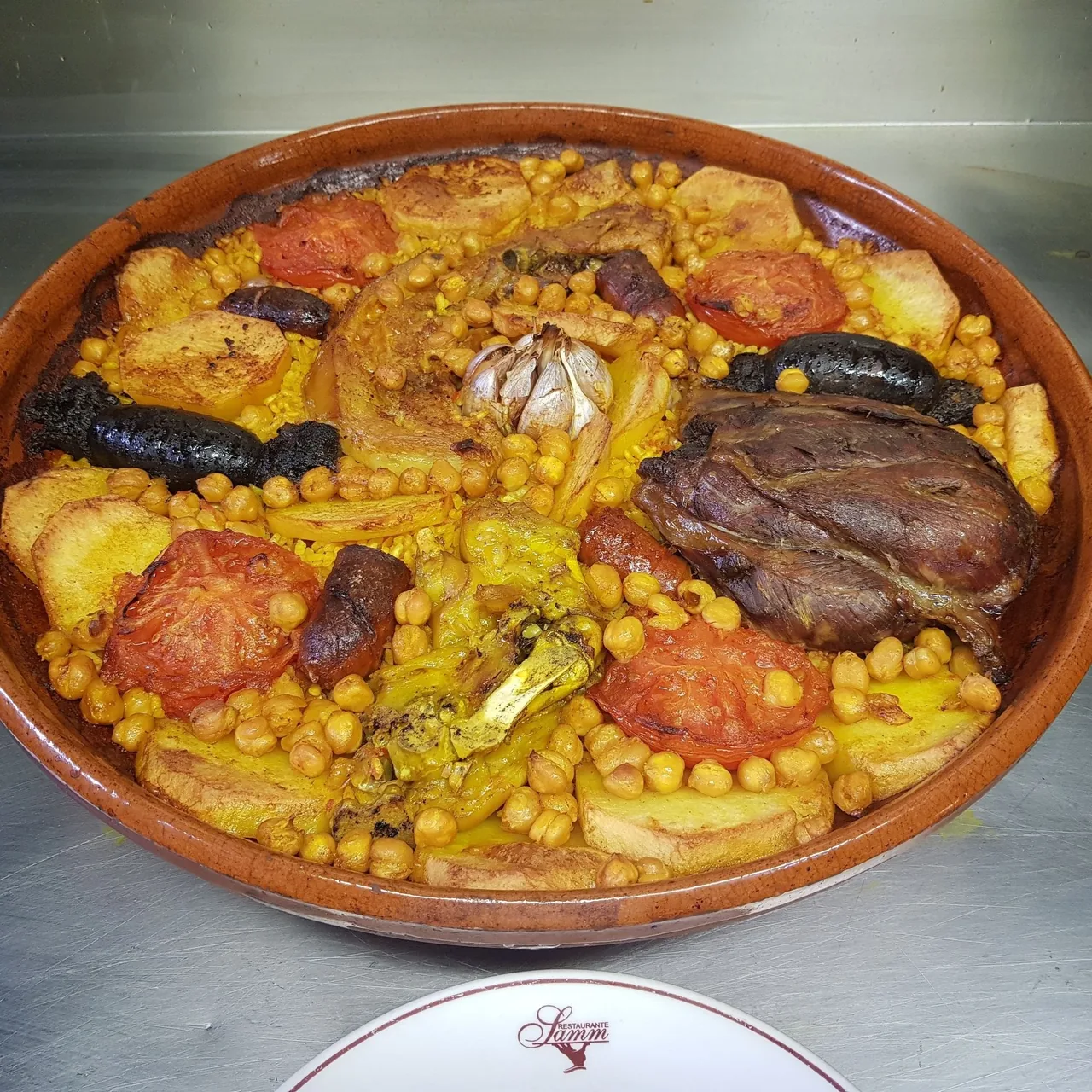 A terracotta dish of baked rice with chickpeas, potato slices, tomato and black pudding