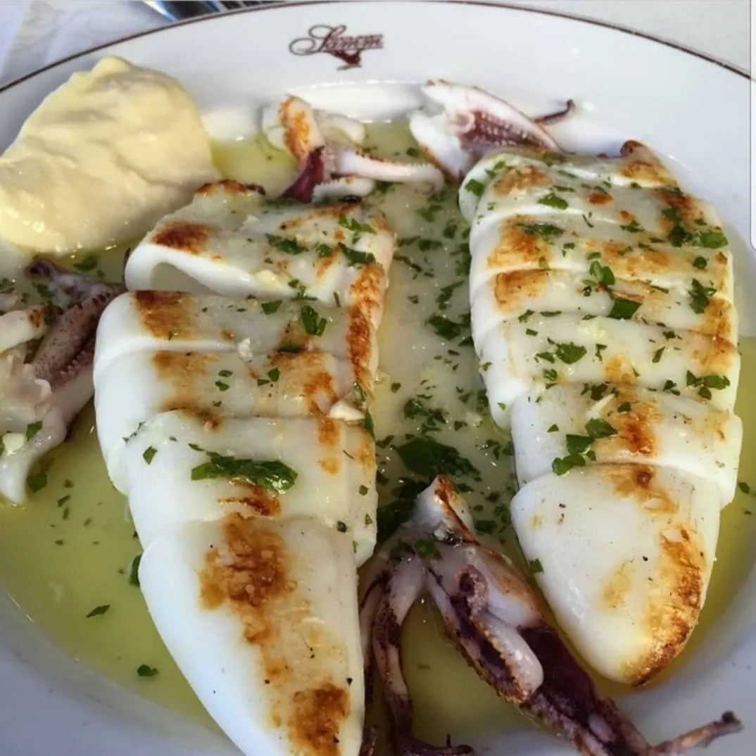 Two grilled squid topped with chopped herbs in olive oil on a white plate