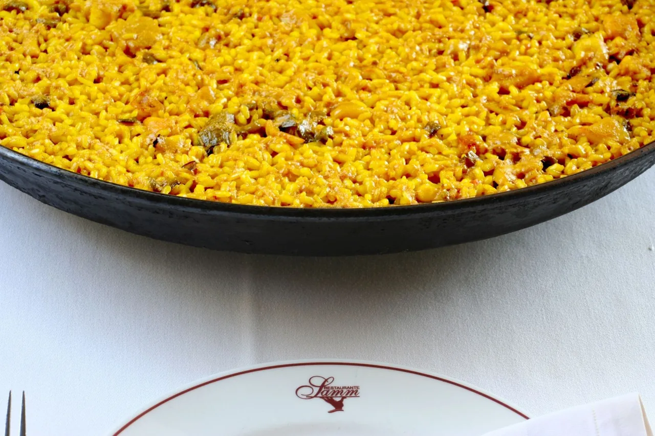 The rim of a paella pan of golden rice beside a Samm-branded plate